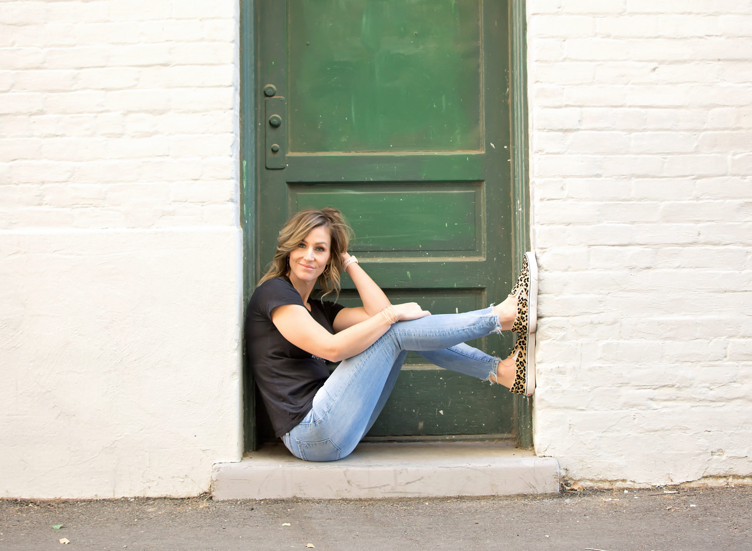 Fitness and Nutrition Coach in a black shirt and blue jeans sitting in front of a green door